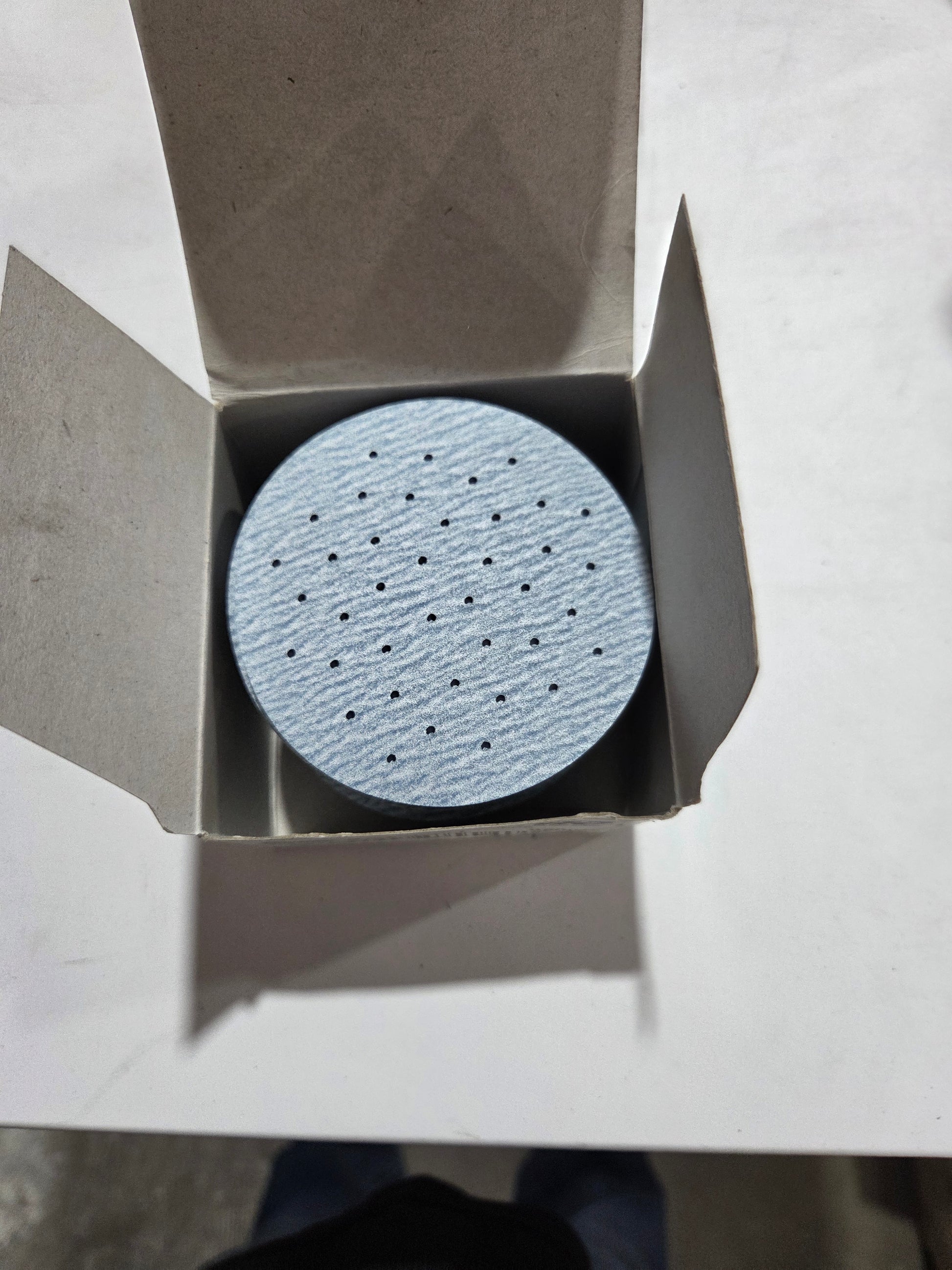 Round sandpaper disc in an open cardboard box on a light gray background