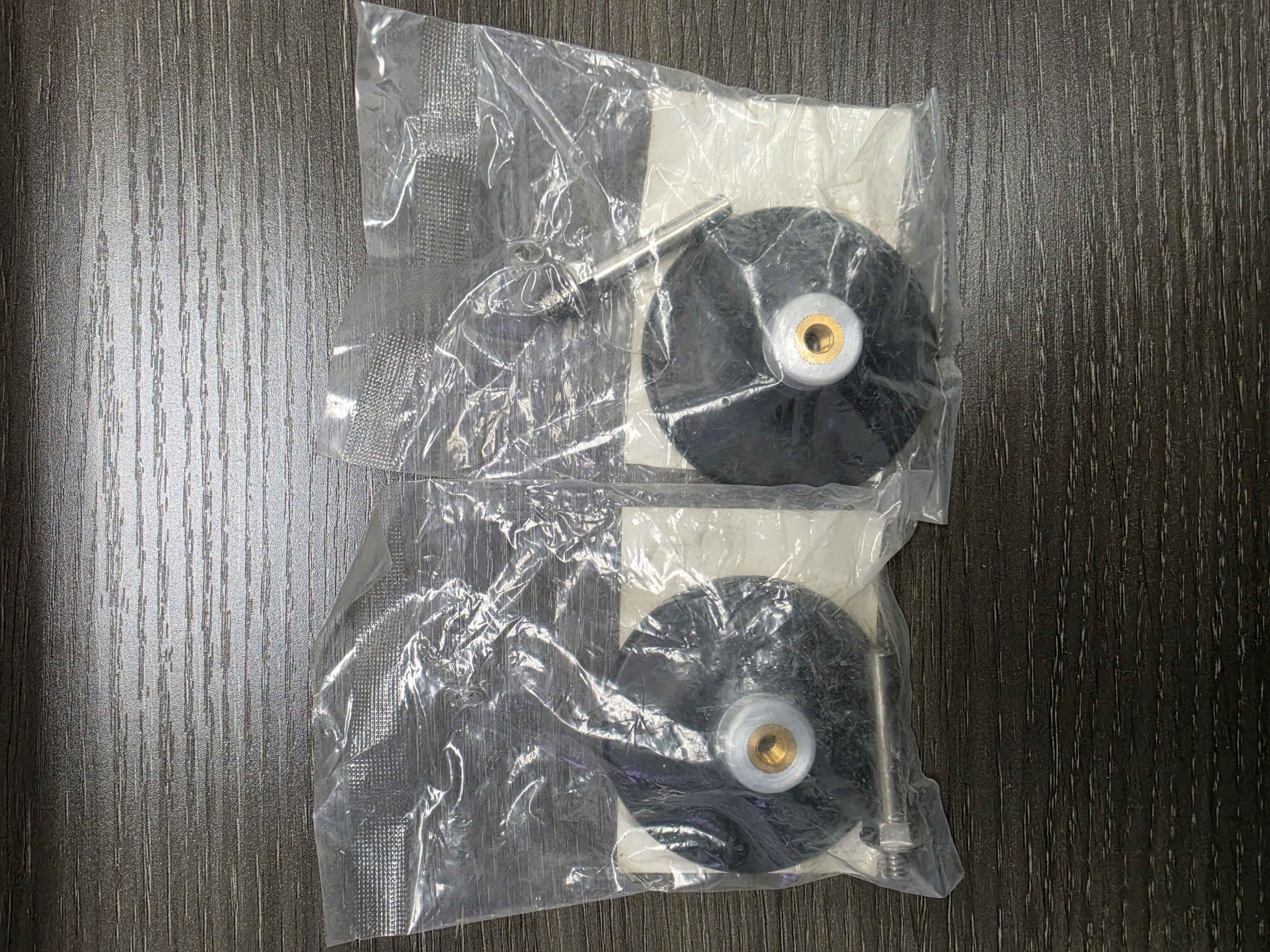 Two black rubber wheels with white axles in a clear plastic bag on a wooden surface.
