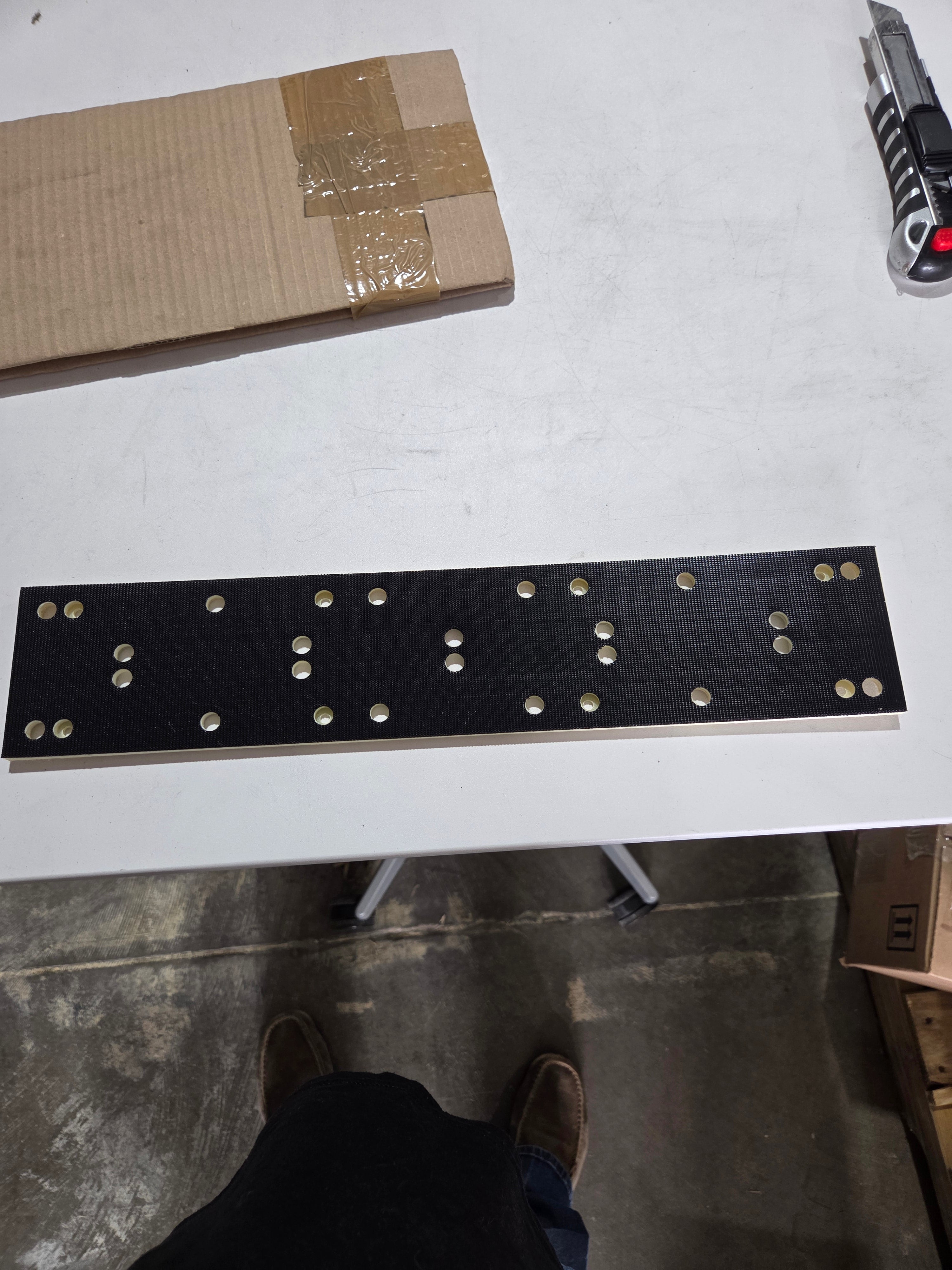 Black board with holes on a white surface, next to cardboard and a tool.