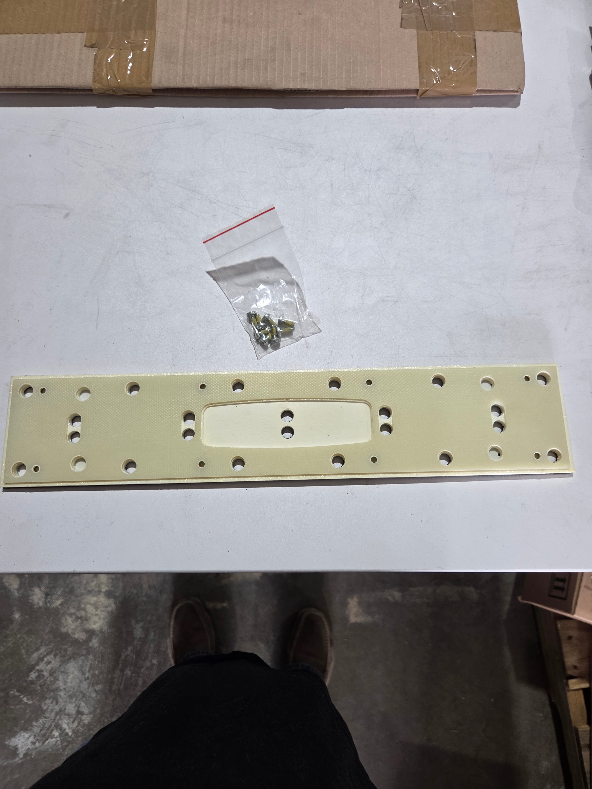 Metal bracket with holes on a white surface, with a small bag of screws nearby.